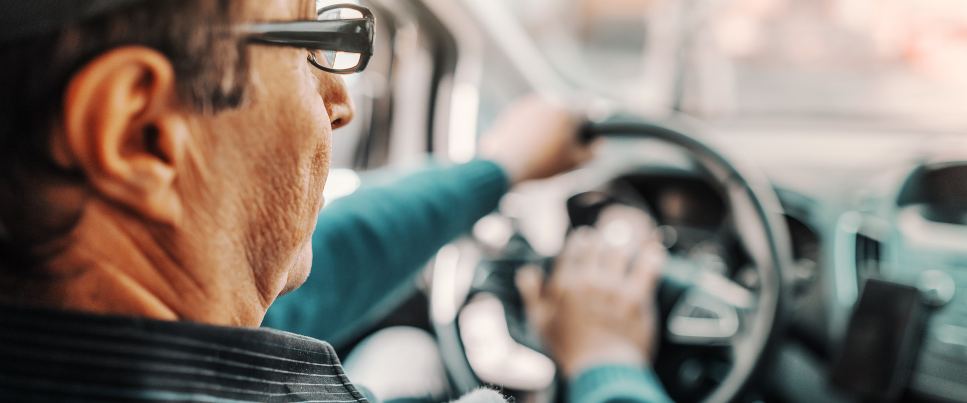 Close-up van een persoon met een bril die een auto bestuurt. Het stuur is zichtbaar en de persoon is gefocust op de weg. Het interieur van de auto is gedeeltelijk zichtbaar, met wazig landschap buiten het raam.