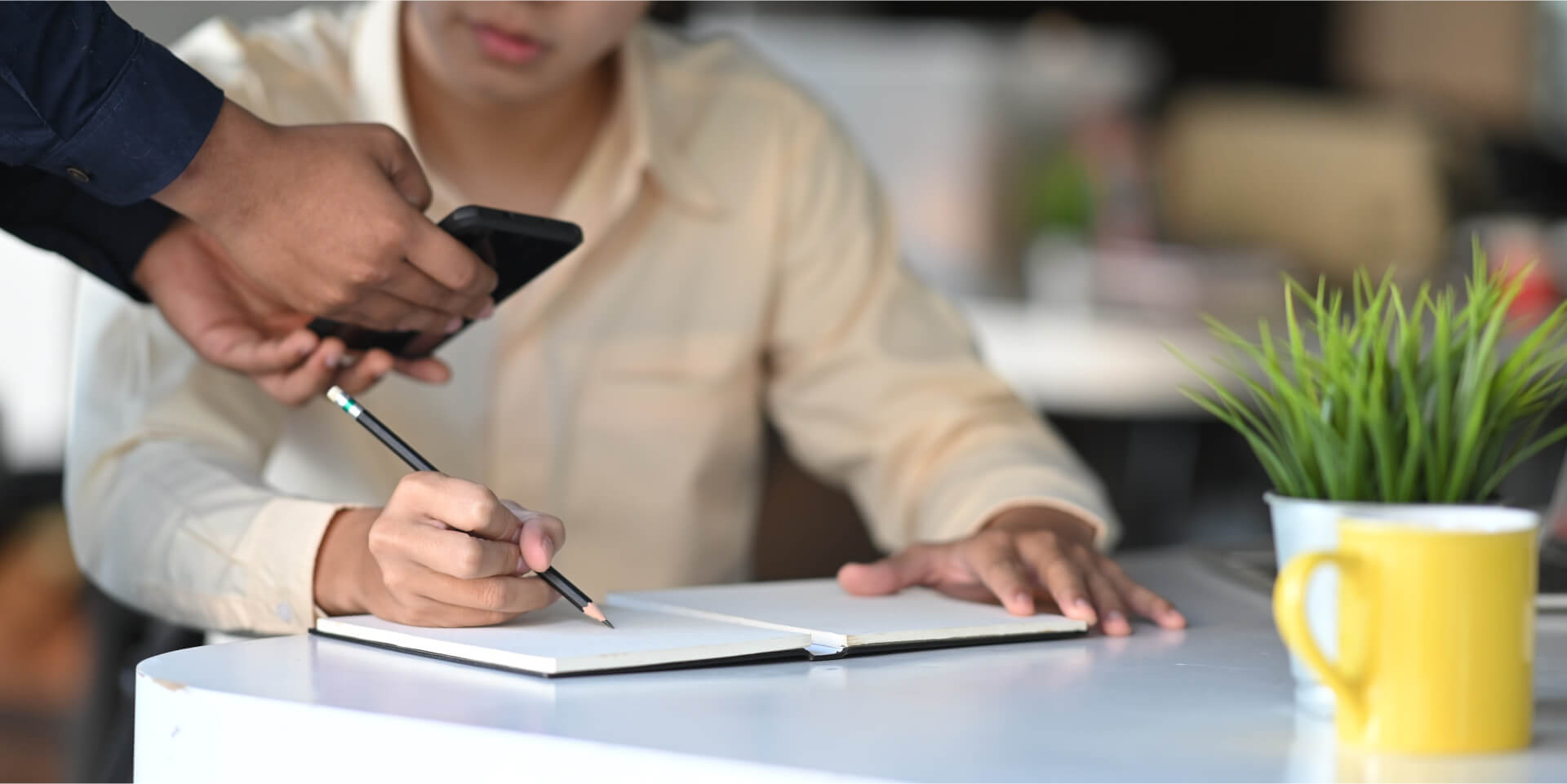 Persoon in een licht shirt schrijft in een notitieboekje aan een bureau. Een andere hand houdt een smartphone boven het notitieboekje. Een gele mok en een groene plant staan op het bureau. De achtergrond is wazig.